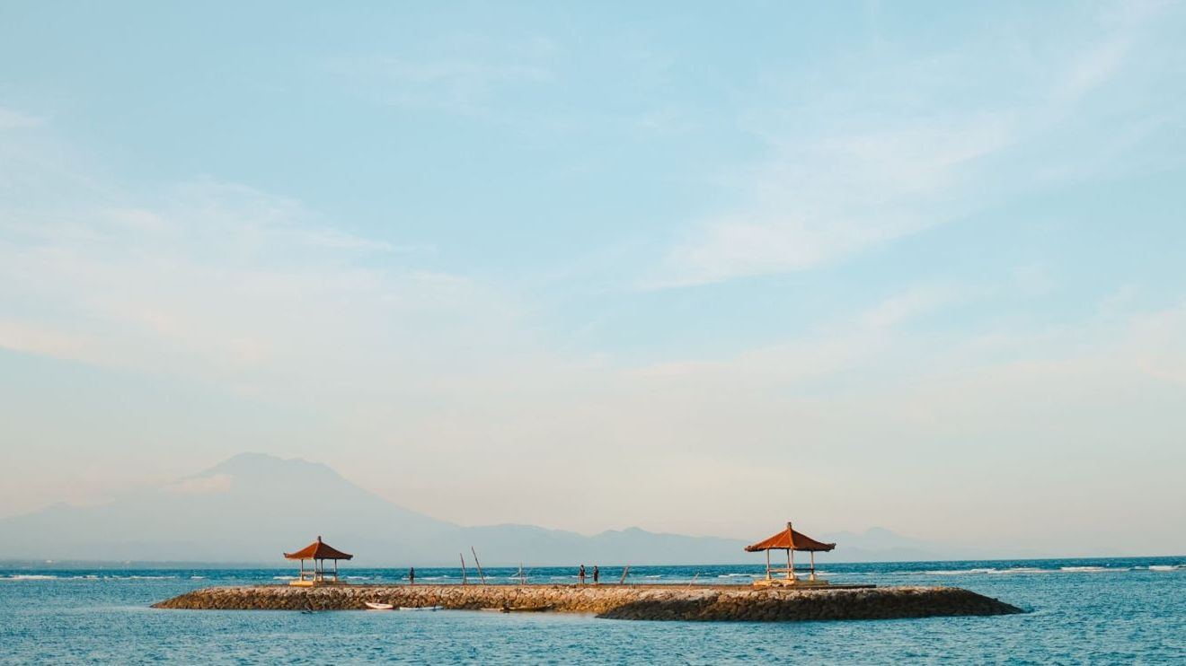 Two Balinese pavilions on a stone jetty off Sanur beach, with Mount Agung faint in the distance
